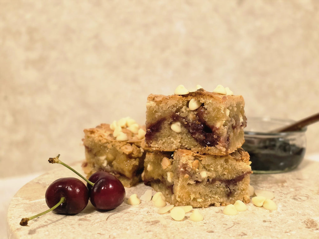 Lady Baker Cherry Bakewell Blondie Mix baked blondies, stacked to show almond sponge, cherry jam swirl, and white chocolate chips, with fresh cherries beside them.