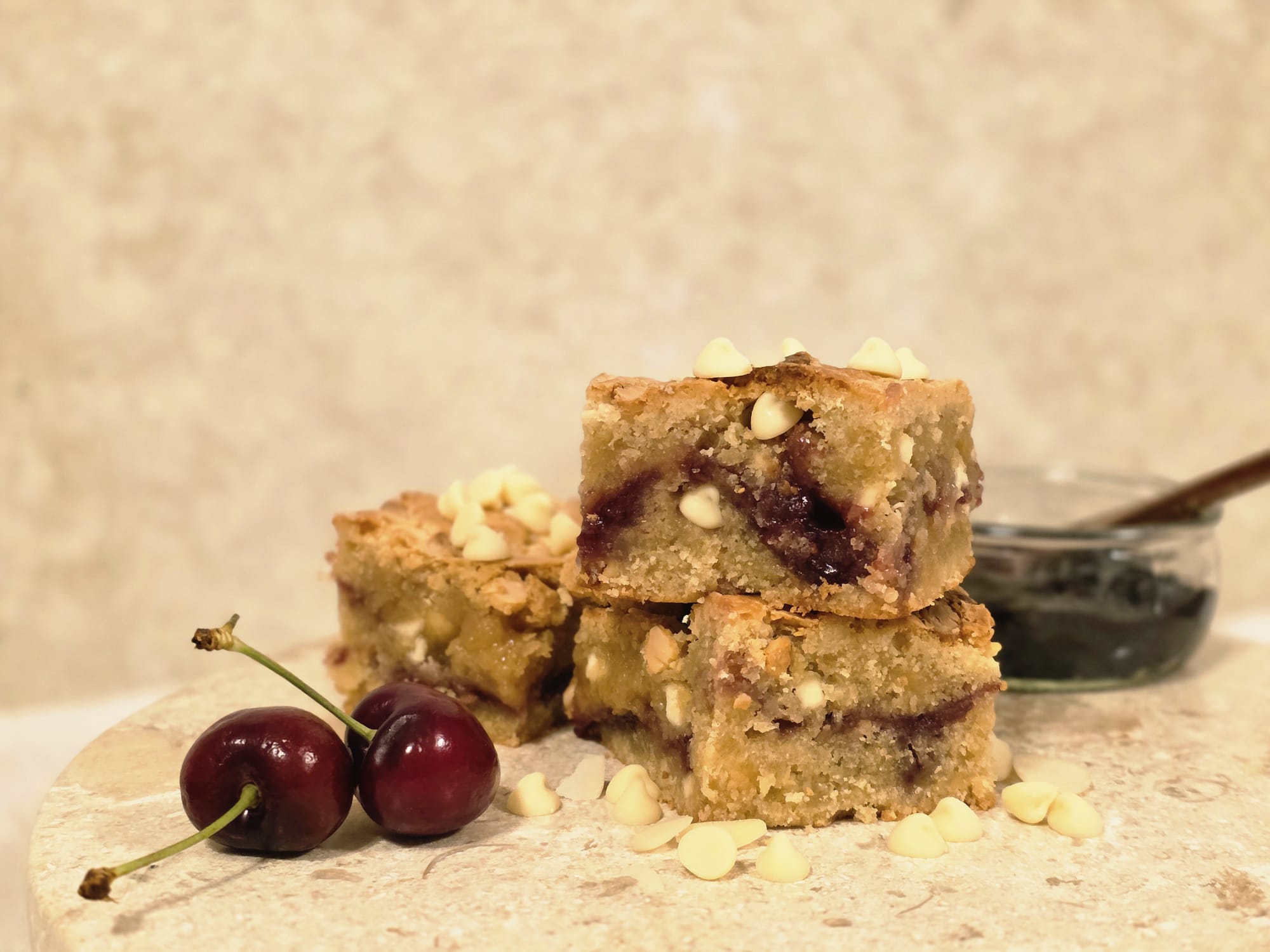 Lady Baker Cherry Bakewell Blondie Mix baked blondies, stacked to show almond sponge, cherry jam swirl, and white chocolate chips, with fresh cherries beside them.