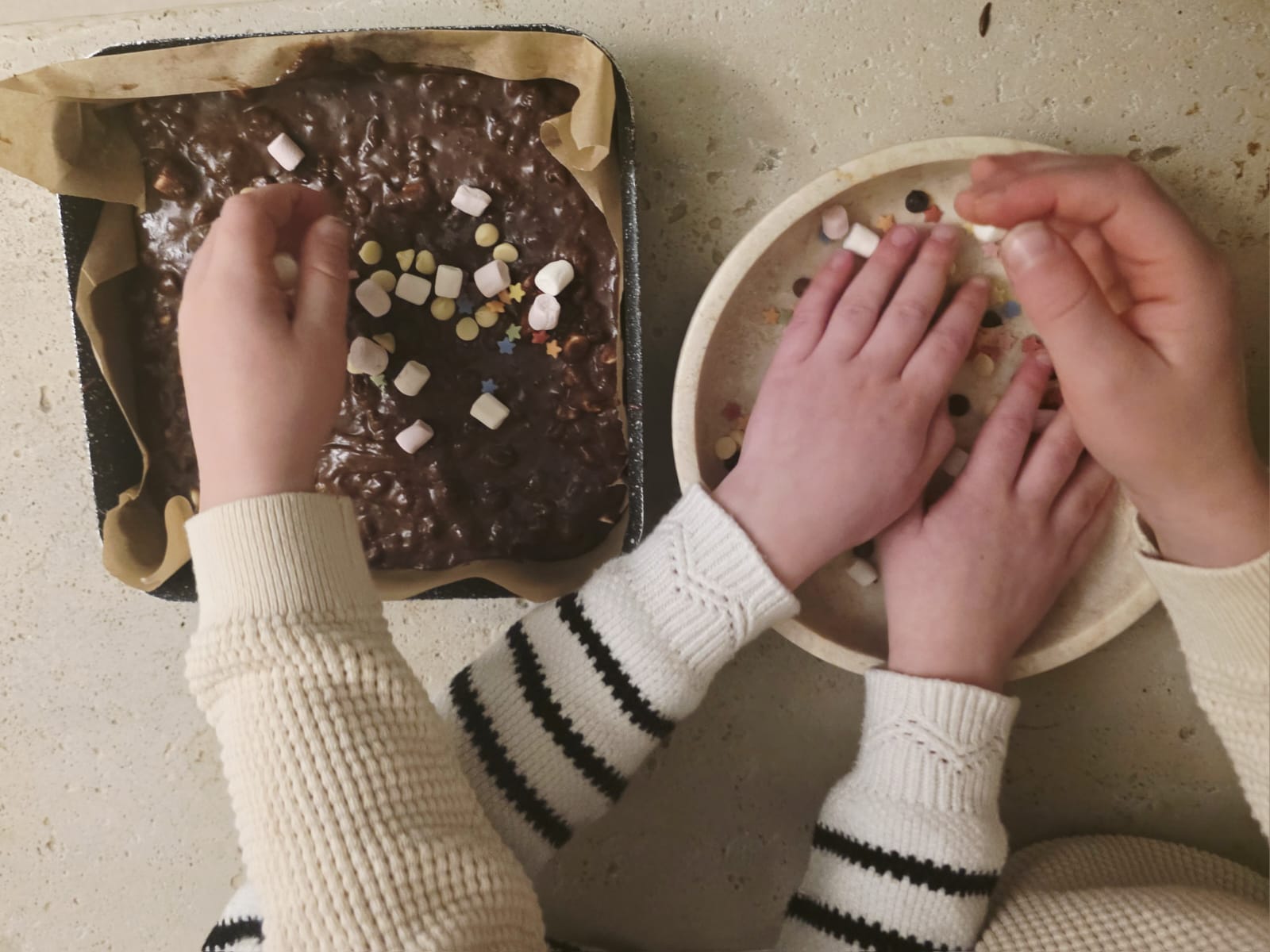 Children decorating a tray of brownie batter with chocolate chips and mini marshmallows from a Lady Baker Magic Brownie Mix.