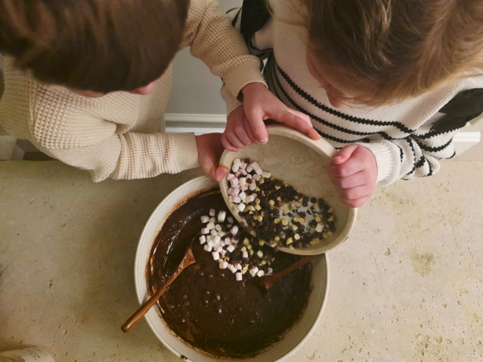 Children pouring chocolate chips and marshmallows from a Lady Baker Magic Brownie Mix jar into a mixing bowl.