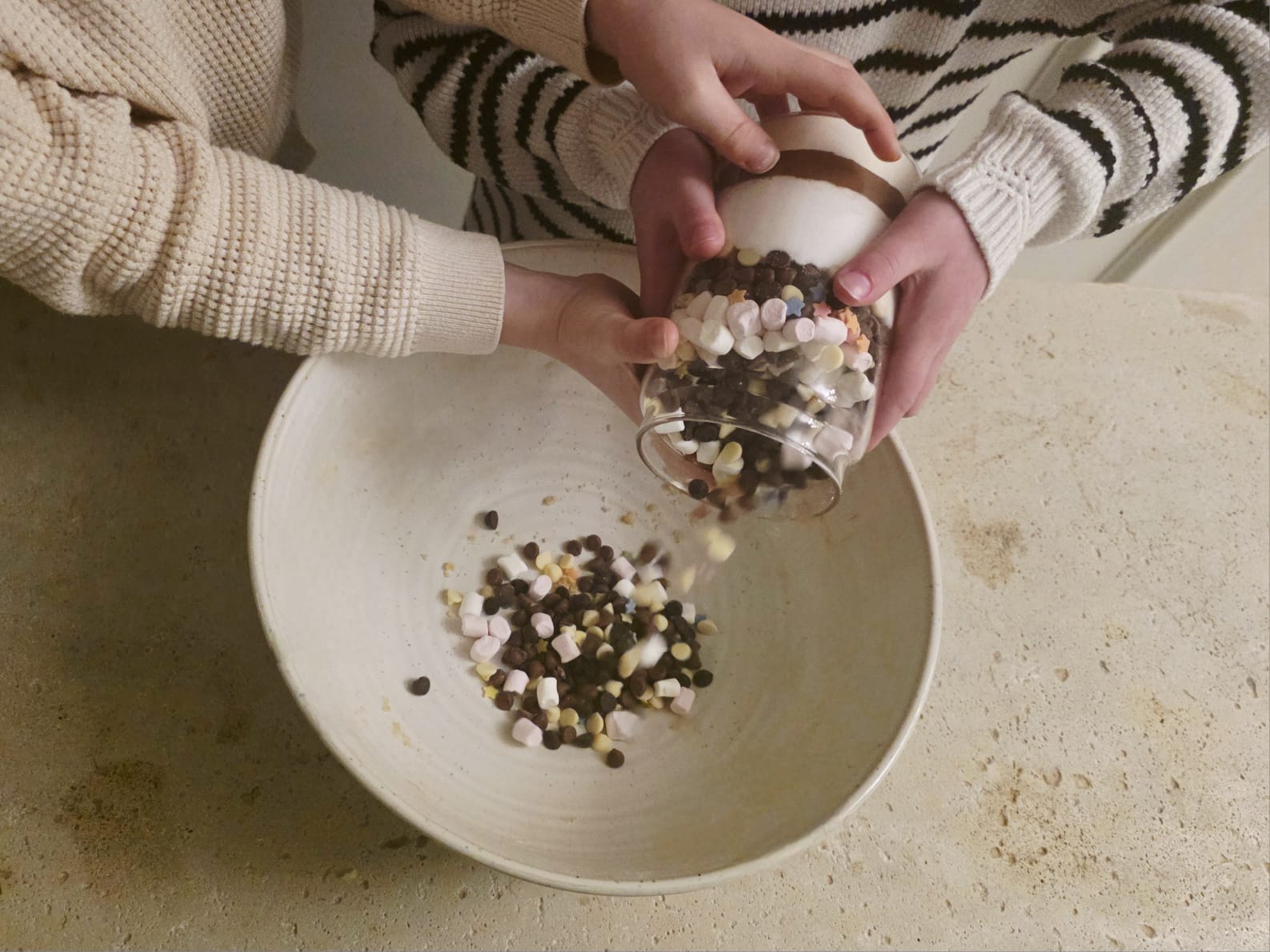 Children pouring chocolate chips and marshmallows from a Lady Baker Magic Brownie Mix jar into a mixing bowl.